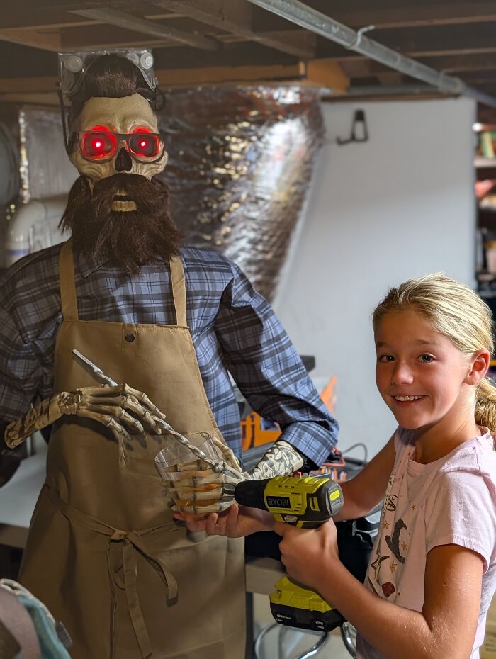 A young girl uses a power drill next to Uncle Murray, a talking skeleton with glowing red eyes. A young girl uses a power drill next to Uncle Murray, a talking skeleton with glowing red eyes.