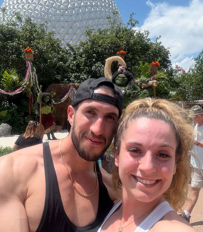 Disney World guest couple taking a selfie outdoors wearing tank tops and caps in the park.