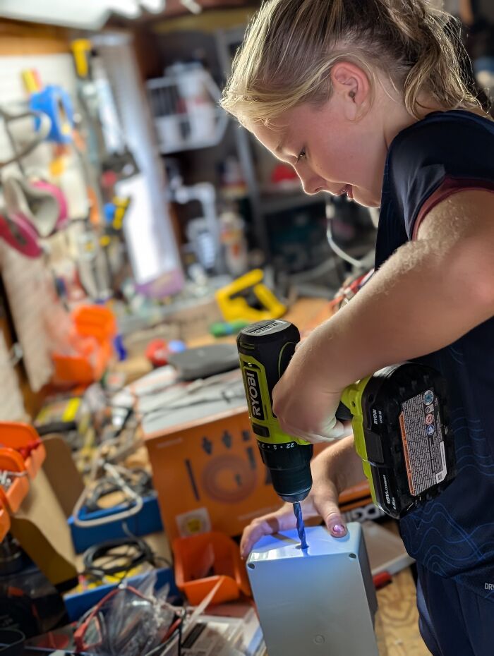A young girl uses a power drill on an electronics enclosure box while working on an AI talking skeleton project. A young girl uses a power drill on an electronics enclosure box while working on an AI talking skeleton project.