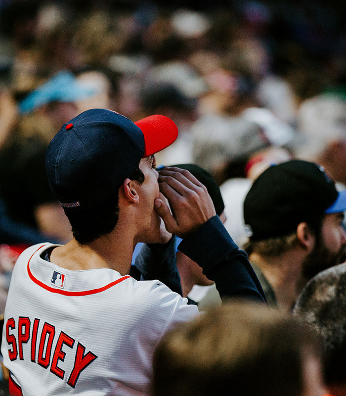 A baseball fan wearing a Spidey jersey shouts from the stands while holding his hand to his mouth.