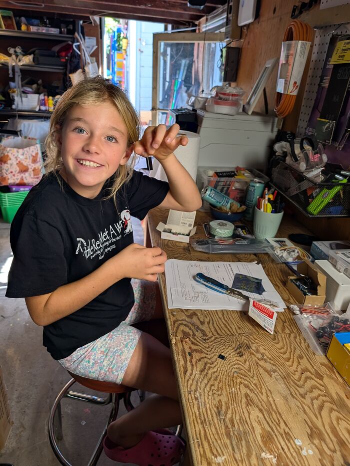 A young girl smiles at her workbench holding a small electronic component for a tiny computer project. A young girl smiles at her workbench holding a small electronic component for a tiny computer project.
