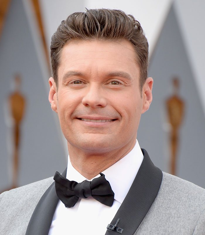 Ryan Seacrest smiling in a grey suit and black bowtie at an event. Ryan Seacrest smiling in a grey suit and black bowtie at an event.