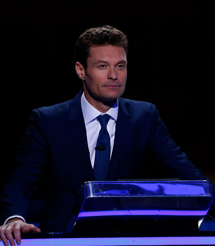 Ryan Seacrest standing behind a podium in a dark suit with blue lighting. Ryan Seacrest standing behind a podium in a dark suit with blue lighting.