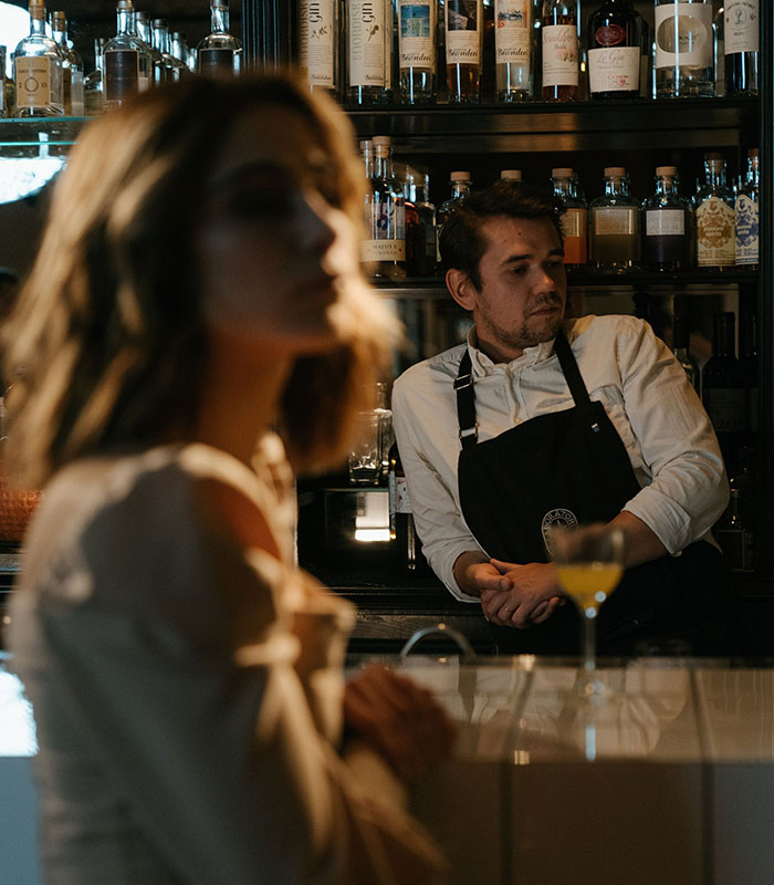 A male bartender standing behind the bar, looking toward a female customer seated in the foreground. A male bartender standing behind the bar, looking toward a female customer seated in the foreground.