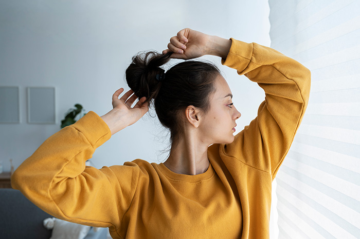 A young woman tying her hair up in a bun indoors, representing a woman's hair hairstyle.