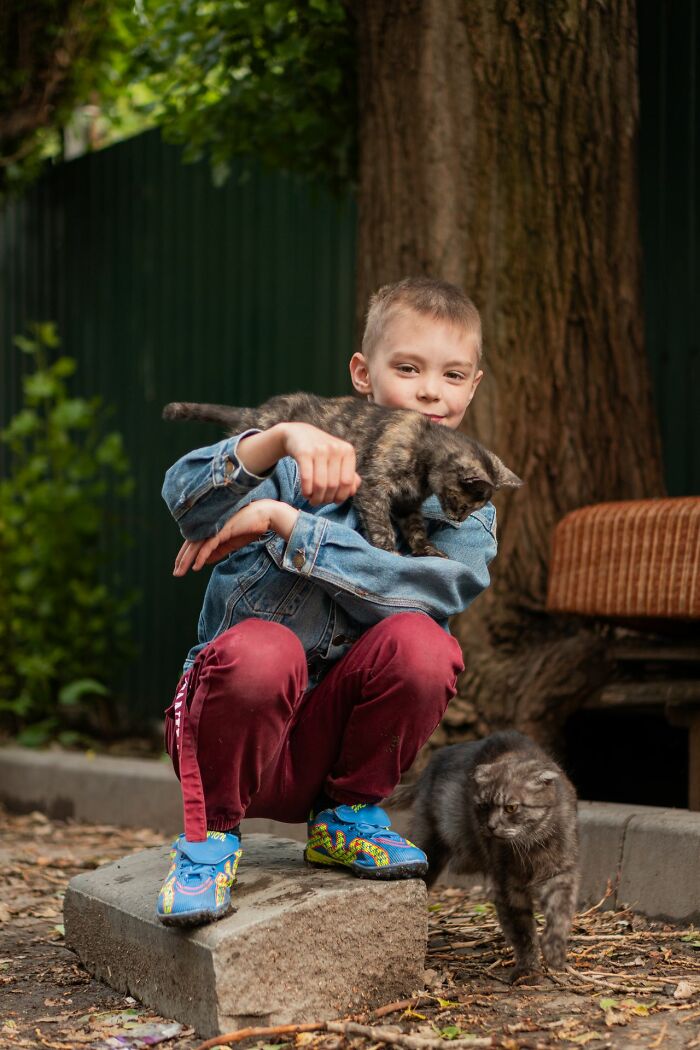 Young boy holding cat while another cat walks nearby in outdoor seriously disturbing scene