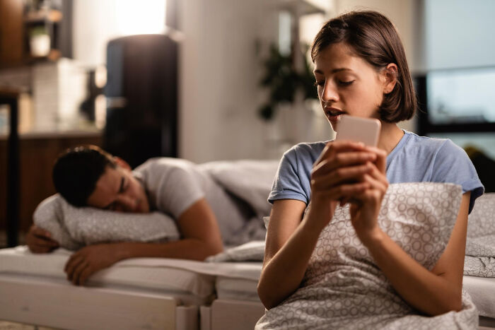 A woman looking at her phone at night while her partner sleeps, considering divorce.