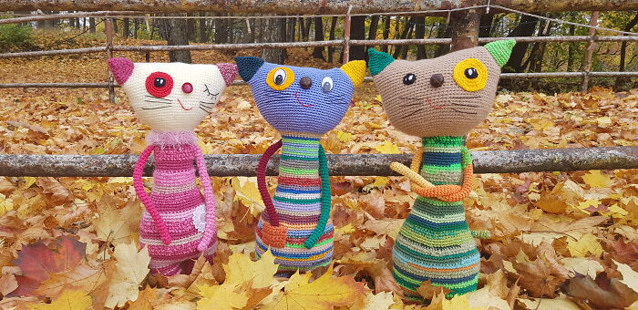 Three colorful crochet creatures shaped like cats sitting on a wooden log among autumn leaves
