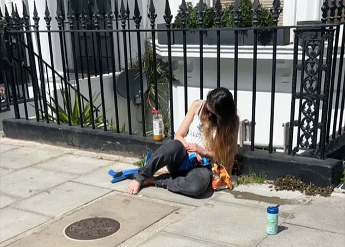 Woman sitting barefoot passed out on London street near black fence