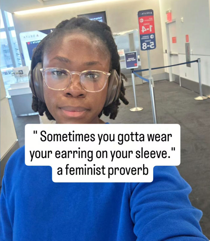 Woman at airport wearing headphones with a quote about wearing earrings on your sleeve feminist proverb Woman at airport wearing headphones with a quote about wearing earrings on your sleeve feminist proverb