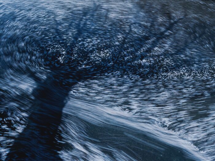 Abstract long exposure landscape photography capturing swirling blue water motion around a tree shadow.