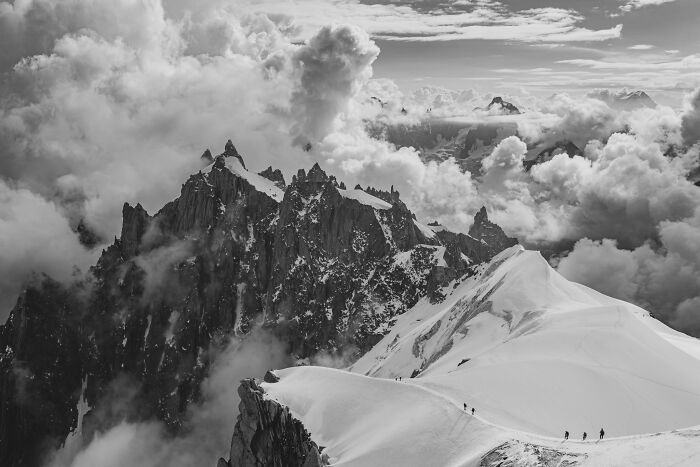 Monochrome landscape photography of jagged snow-covered mountain peaks under dramatic clouds.