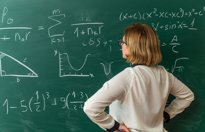 A female teacher wearing glasses stands with hands on hips looking at complex math formulas on a blackboard