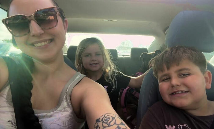 A mom taking a selfie in a car with her 2 children smiling in the back and passenger seats.