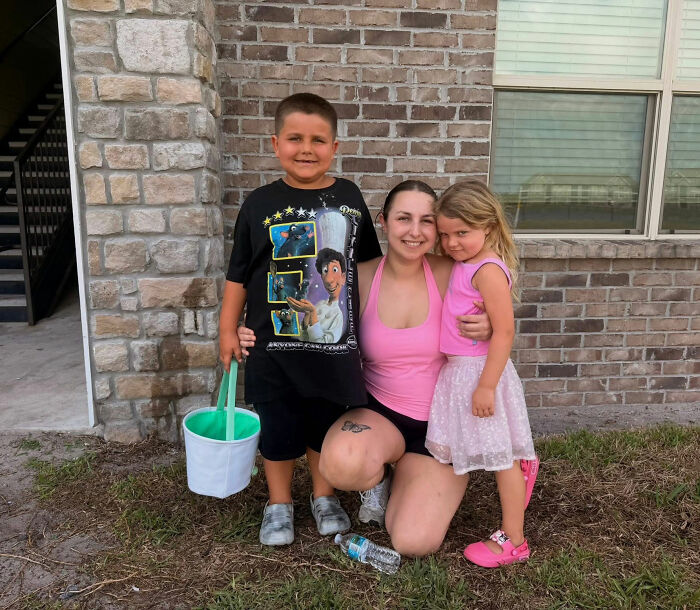 A smiling mom posing outside with her 2 children, holding hands near a brick building.