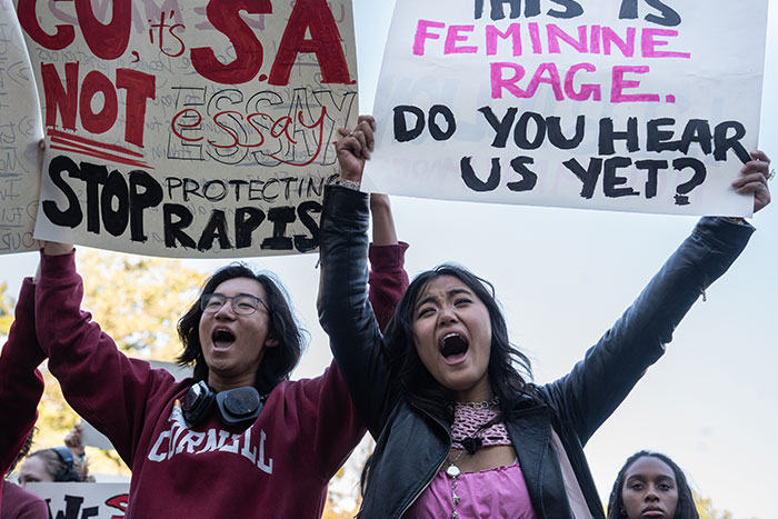 Protesters shouting while holding signs against s****l a*****t related to the Cornell 7 scandal.