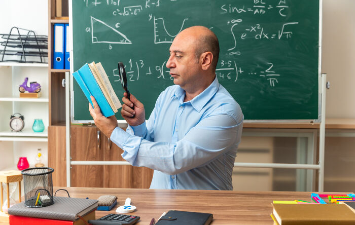 A male teacher looking closely at a book through a magnifying glass in front of a green chalkboard
