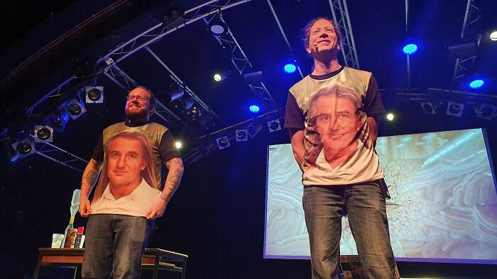Two men on a stage wearing printed face shirts, performing in front of an audience about unusual cults