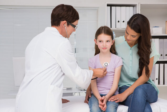 Doctor using stethoscope on young girl sitting with mother during checkup