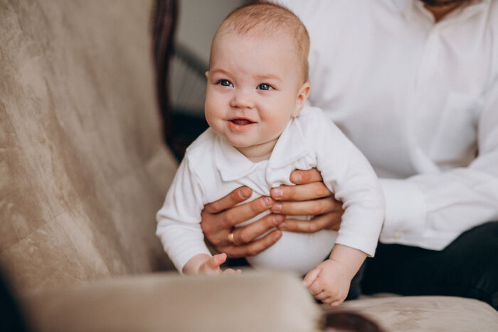 An adult holding a cute smiling baby dressed in white on a sofa.
