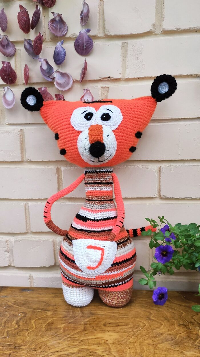 A funny orange striped tiger crochet creature with big cartoon eyes standing against a brick wall
