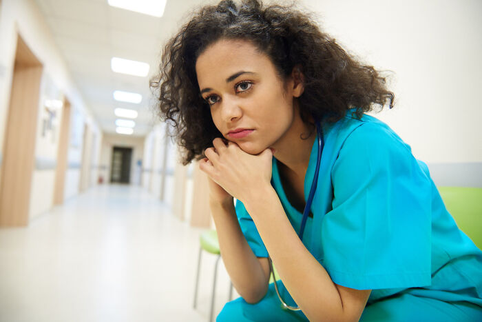 A thoughtful female healthcare worker in teal scrubs sits in a hospital corridor.
