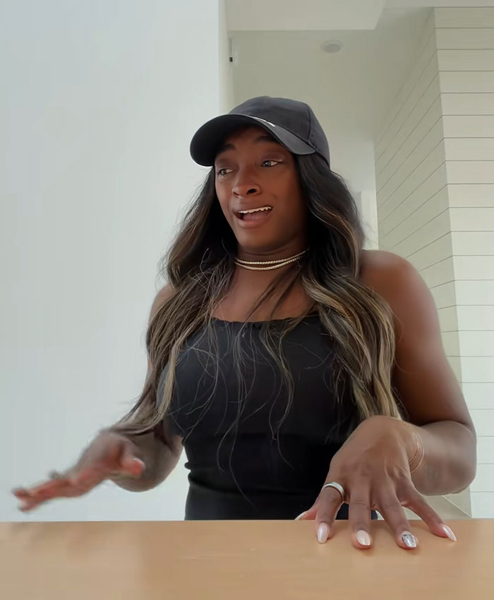 Simone Biles expressing emotion in a cap and black tank top while discussing selling her Texas home