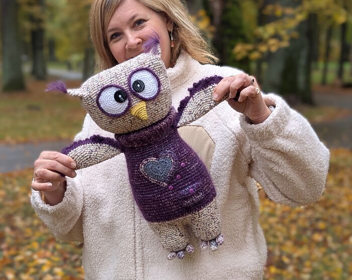 A smiling woman holding a large, handmade crochet creature owl wearing a purple sweater in an autumn park