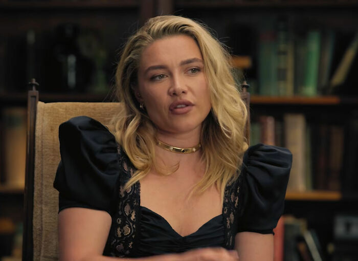 Florence Pugh with blonde hair looking thoughtful while sitting in front of a bookshelf.