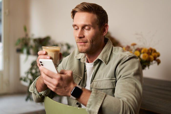 A man holding a coffee cup and looking at his smartphone, reading emotional divorce stories online.