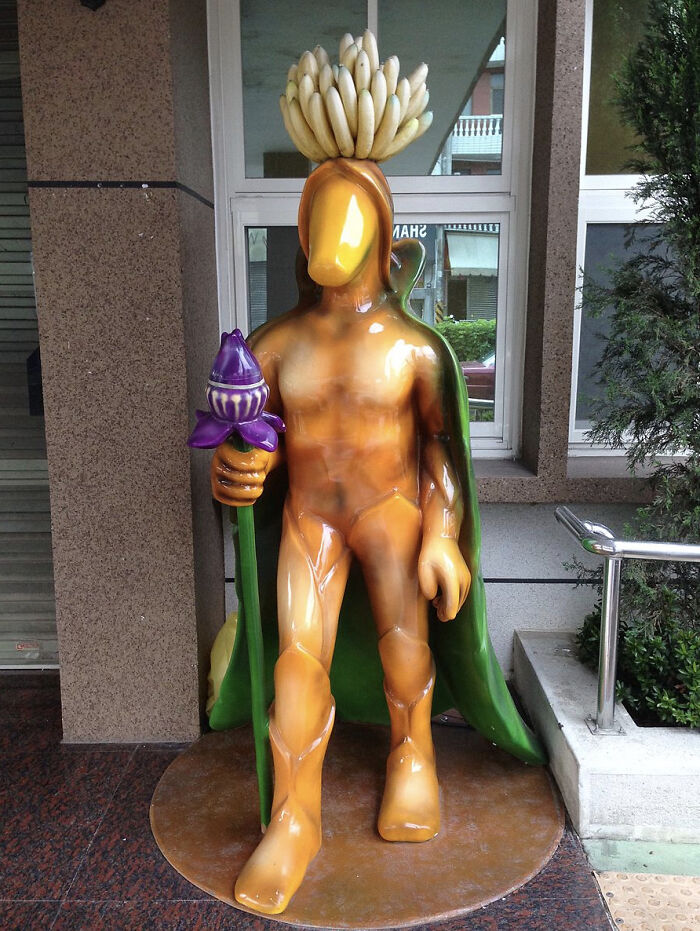 A bizarre orange human-shaped statue of public art featuring bananas on its head and holding a purple bud