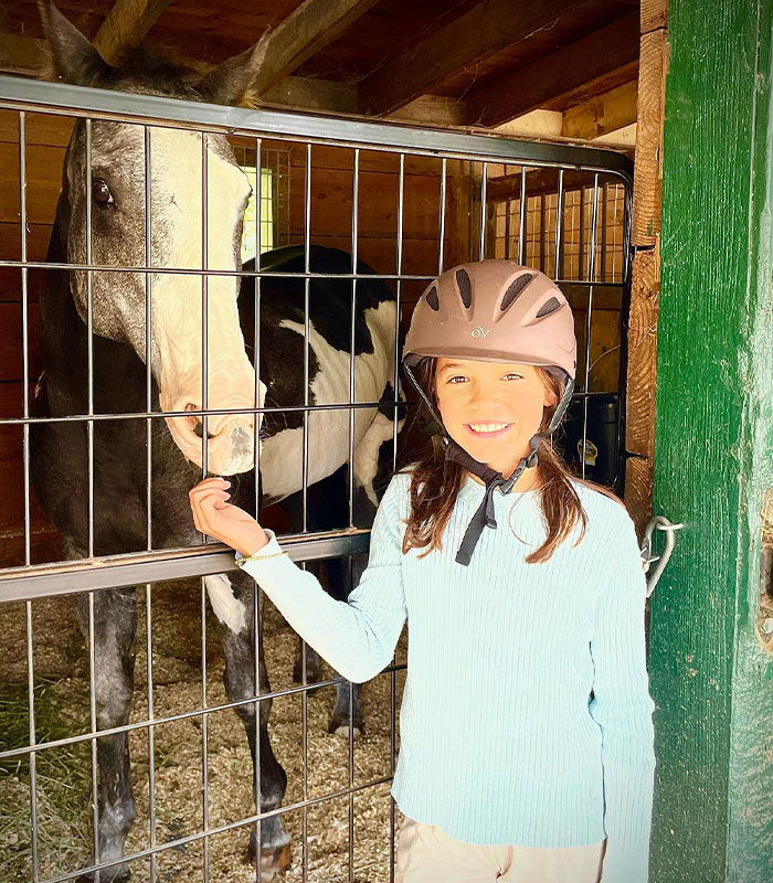Fiona Lowe, daughter of Chad Lowe, smiles wearing a riding helmet next to a horse in a stable.