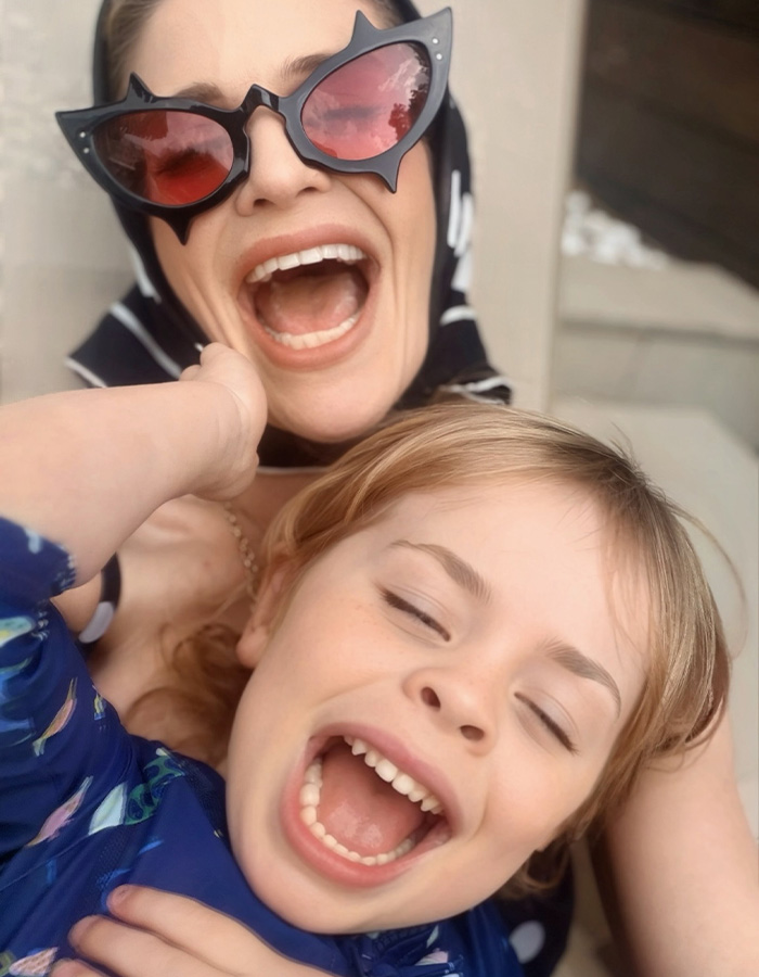Kelly Osbourne laughing outdoors with her young child wearing oversized bat-wing sunglasses.