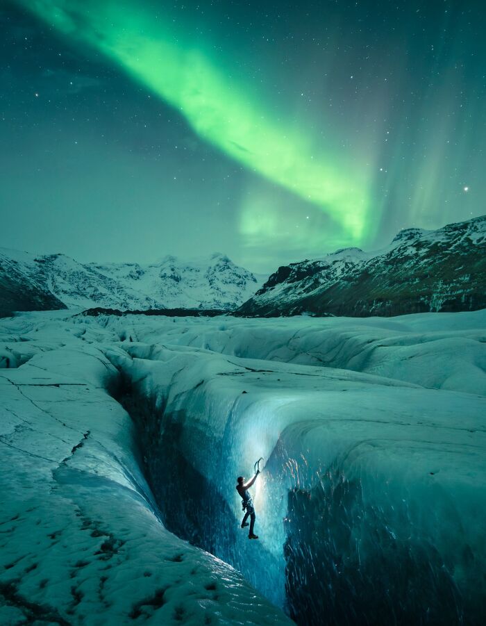 Natural landscape photography of a climber inside a glowing ice crevasse under northern lights.