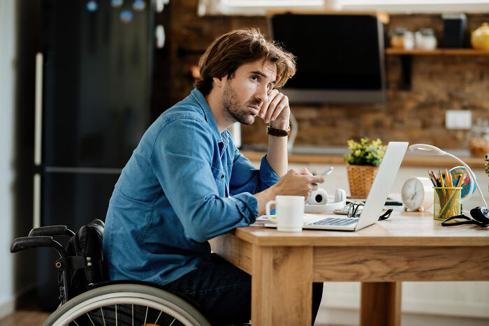 A pensive man in a wheelchair looking away while sitting at a desk with a laptop, reflecting divorce stories.