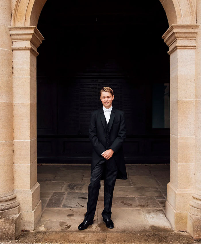 Prince George smiles while wearing a formal black suit and standing under a stone archway. Prince George smiles while wearing a formal black suit and standing under a stone archway.