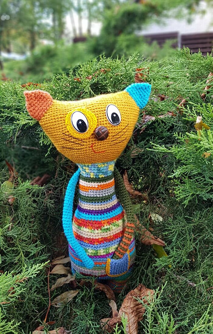 A yellow crochet cat toy with striped body stands nestled in green evergreen bushes.