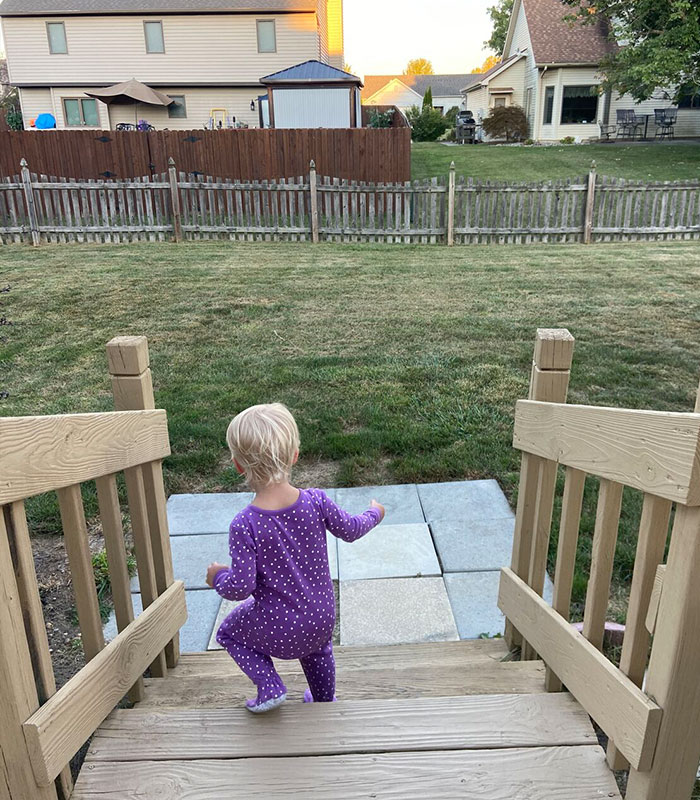 A toddler in purple pajamas walking down wooden outdoor steps into a backyard.
