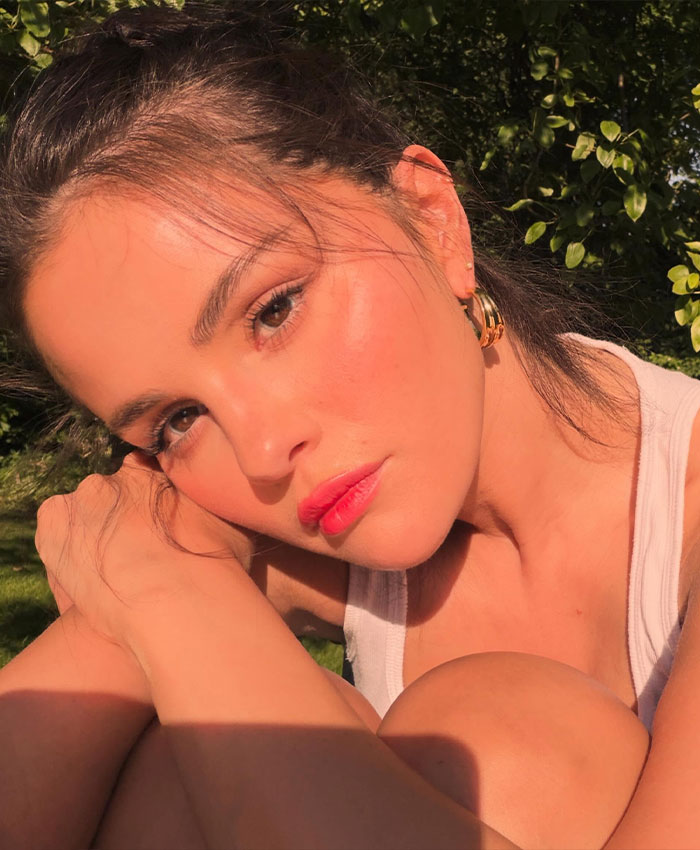 Selena Gomez taking a sunlit close-up selfie outdoors while resting her head on her knees.