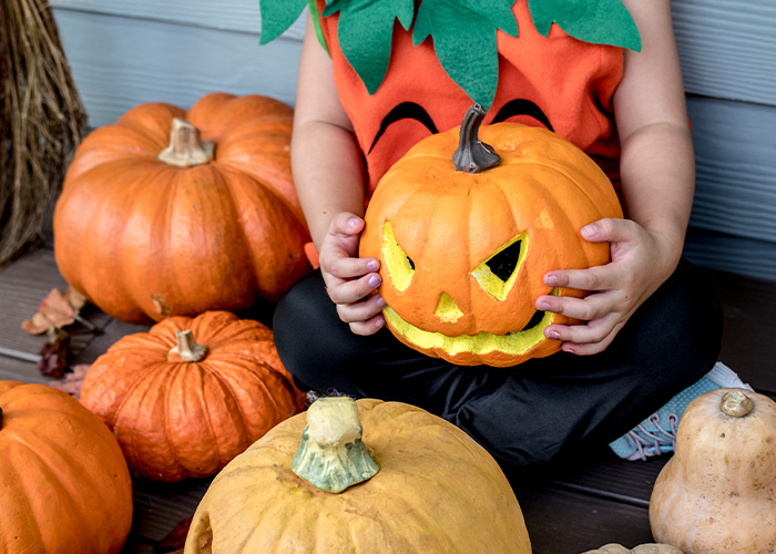 A toddler dressed in a pumpkin costume holding a carved jack-o-lantern during a Halloween activity.