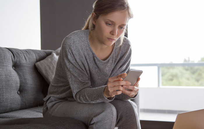Concerned woman sitting on couch looking at smartphone while boyfriend gets heart surgery.