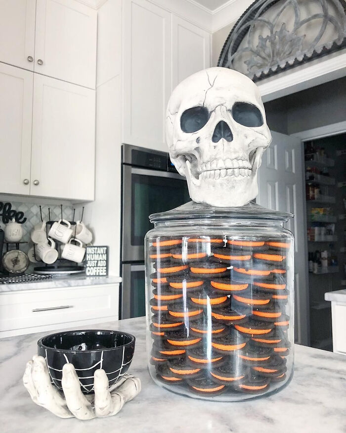 Halloween skull decoration sitting atop a large glass jar filled with black and orange cookies.