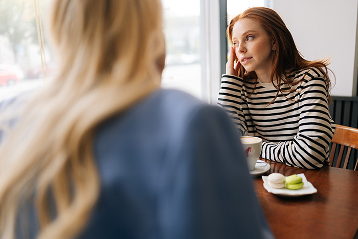 Two women sitting at a cafe table, with one woman uncovering layer after layer of lies before her ex's funeral. Two women sitting at a cafe table, with one woman uncovering layer after layer of lies before her ex's funeral.
