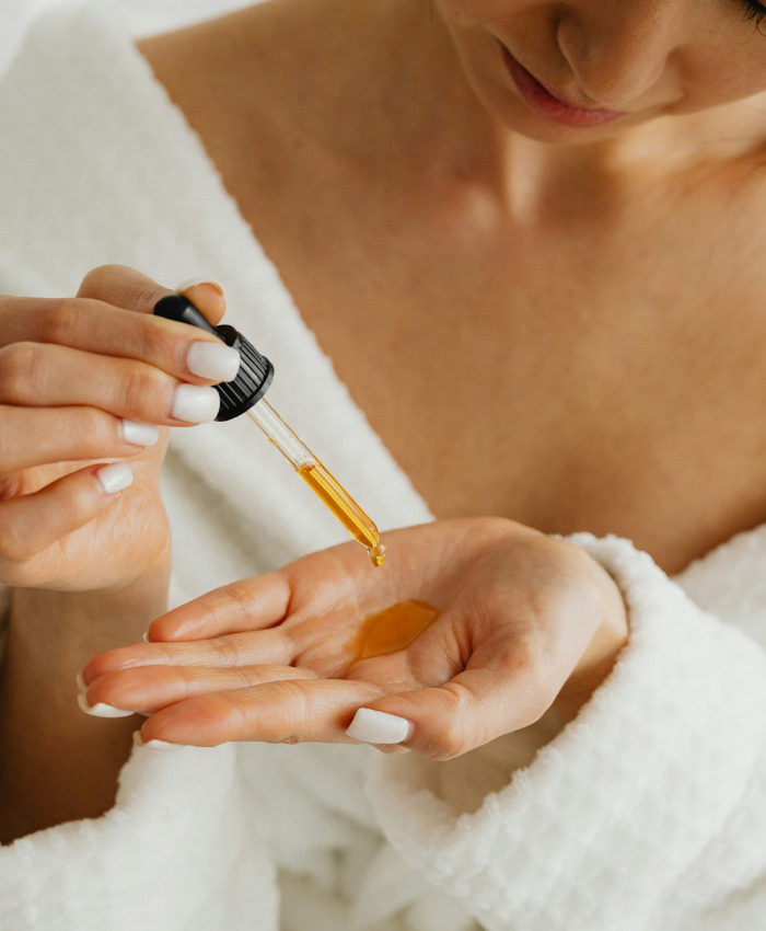 Woman in a white robe using a dropper to apply golden facial oil onto her hand Woman in a white robe using a dropper to apply golden facial oil onto her hand