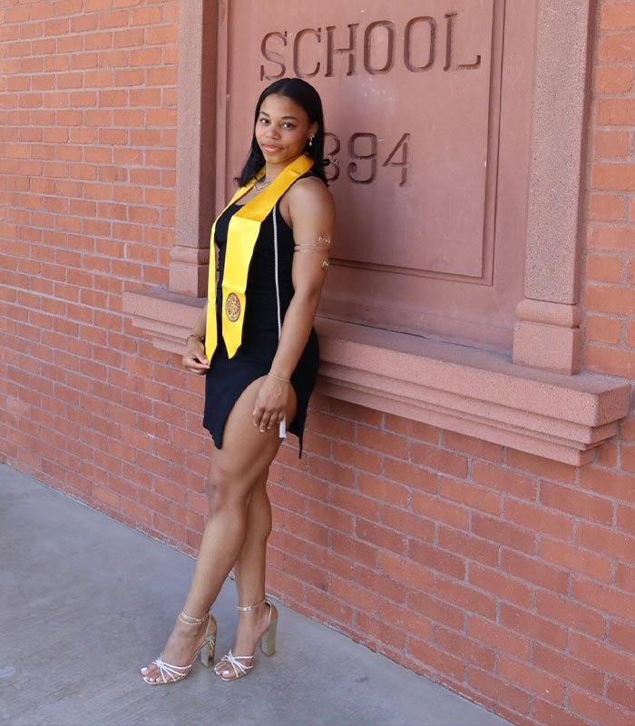 Arizona State Cheerleader posing in a black dress with a graduation sash by a school wall