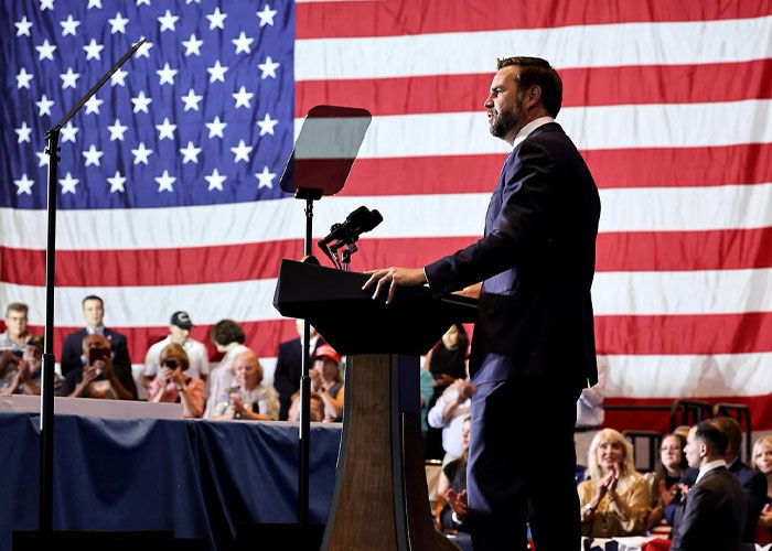 JD Vance gesturing while delivering a speech at a podium with a large American flag behind him. JD Vance gesturing while delivering a speech at a podium with a large American flag behind him.