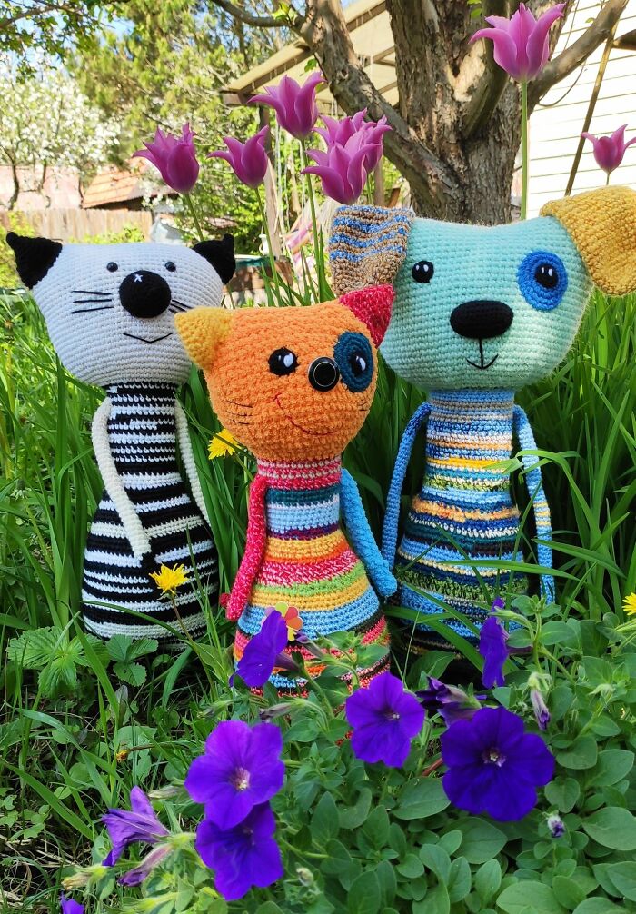 Three colorful handmade crochet creatures stand together in a grassy garden near purple flowers.