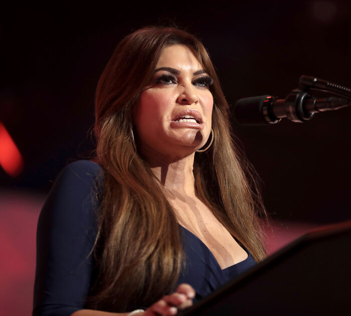MAGA influencer speaking at microphone with long brown hair and dark dress