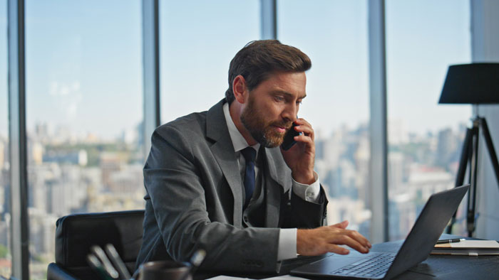 A professional businessman talking on his phone while typing on his laptop in a high-rise office