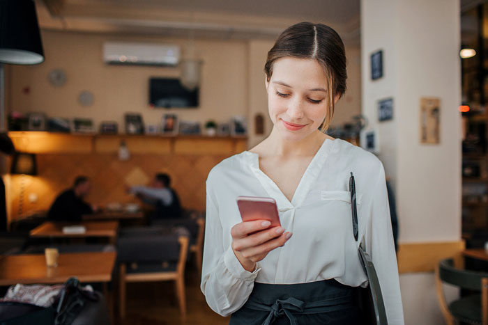 A smiling waitress in a cafe holding a phone, symbolizing a bossy coworker messaging managers about a waiter A smiling waitress in a cafe holding a phone, symbolizing a bossy coworker messaging managers about a waiter